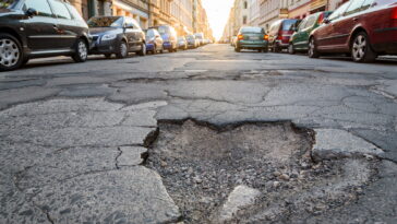Grande buca sull’asfalto in una strada urbana con auto parcheggiate ai lati.