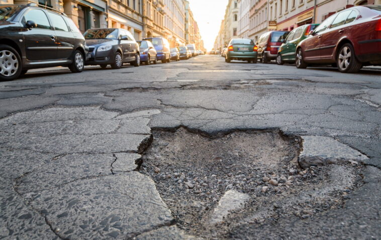Grande buca sull’asfalto in una strada urbana con auto parcheggiate ai lati.