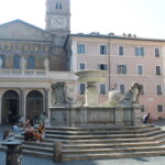 La storica fontana di Piazza Santa Maria in Trastevere, con la basilica sullo sfondo e persone sedute sui gradini in una giornata di sole.