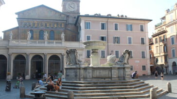 La storica fontana di Piazza Santa Maria in Trastevere, con la basilica sullo sfondo e persone sedute sui gradini in una giornata di sole.
