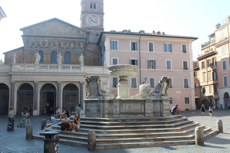 La storica fontana di Piazza Santa Maria in Trastevere, con la basilica sullo sfondo e persone sedute sui gradini in una giornata di sole.
