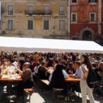 Persone sedute ai tavoli durante una sagra di paese nel Lazio con stand gastronomici e bancarelle.