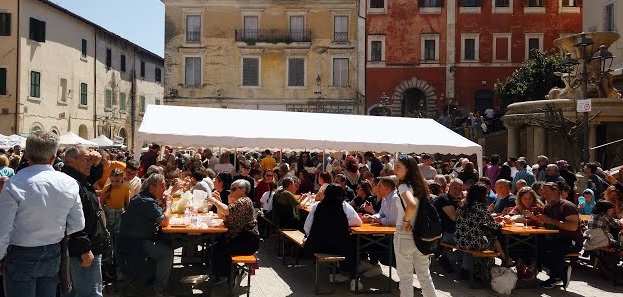 Persone sedute ai tavoli durante una sagra di paese nel Lazio con stand gastronomici e bancarelle.