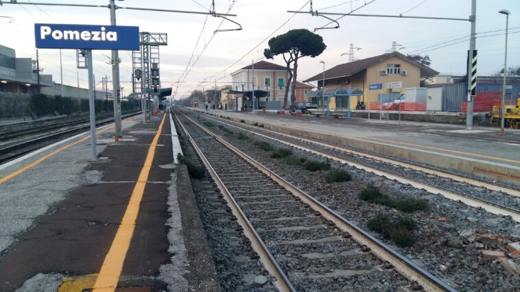 Binari e banchina della stazione di Pomezia con cartello visibile e linee ferroviarie.