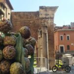 Carciofi romaneschi esposti in primo piano in una strada di Roma con edifici storici e persone che passeggiano sullo sfondo.