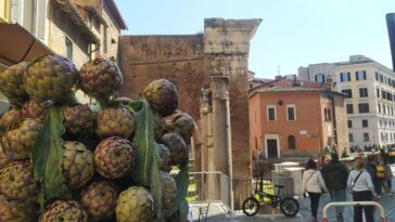 Carciofi romaneschi esposti in primo piano in una strada di Roma con edifici storici e persone che passeggiano sullo sfondo.