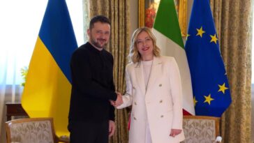Volodymyr Zelensky stringe la mano a Giorgia Meloni durante un incontro istituzionale, con le bandiere di Ucraina, Italia e Unione Europea sullo sfondo.