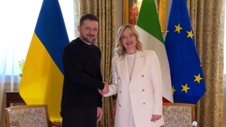 Volodymyr Zelensky stringe la mano a Giorgia Meloni durante un incontro istituzionale, con le bandiere di Ucraina, Italia e Unione Europea sullo sfondo.
