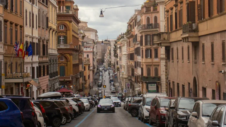 Strada di Roma con traffico e auto parcheggiate lungo i lati, palazzi storici ai bordi