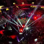 Vista dall’alto di un grande concerto indoor con migliaia di spettatori, luci rosse e bianche che illuminano il palco centrale durante uno spettacolo musicale.
