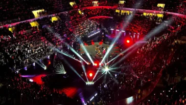 Vista dall’alto di un grande concerto indoor con migliaia di spettatori, luci rosse e bianche che illuminano il palco centrale durante uno spettacolo musicale.