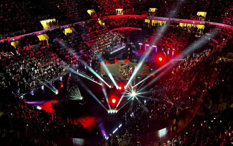 Vista dall’alto di un grande concerto indoor con migliaia di spettatori, luci rosse e bianche che illuminano il palco centrale durante uno spettacolo musicale.