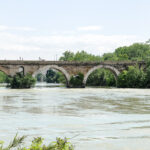 Antico ponte in pietra sul fiume Tevere circondato dal verde, con acqua che scorre sotto le arcate e vegetazione rigogliosa lungo le sponde.