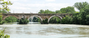 Antico ponte in pietra sul fiume Tevere circondato dal verde, con acqua che scorre sotto le arcate e vegetazione rigogliosa lungo le sponde.