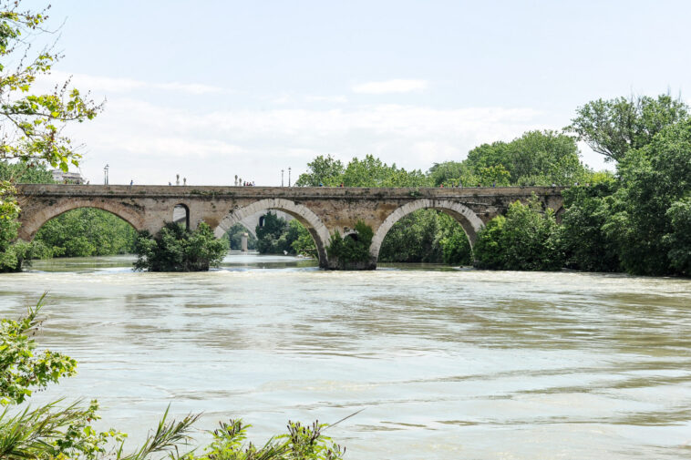 Antico ponte in pietra sul fiume Tevere circondato dal verde, con acqua che scorre sotto le arcate e vegetazione rigogliosa lungo le sponde.