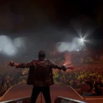 Vasco Rossi sul palco durante un grande concerto davanti a decine di migliaia di fan. Il cantante, visto di spalle con le braccia aperte, domina una folla illuminata dalle luci e dagli smartphone accesi.
