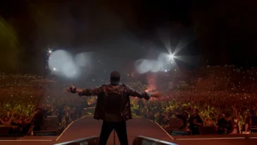 Vasco Rossi sul palco durante un grande concerto davanti a decine di migliaia di fan. Il cantante, visto di spalle con le braccia aperte, domina una folla illuminata dalle luci e dagli smartphone accesi.