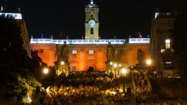 Piazza del Campidoglio illuminata durante un evento serale a Roma, con centinaia di persone riunite sulla scalinata e nella piazza davanti al Palazzo Senatorio.