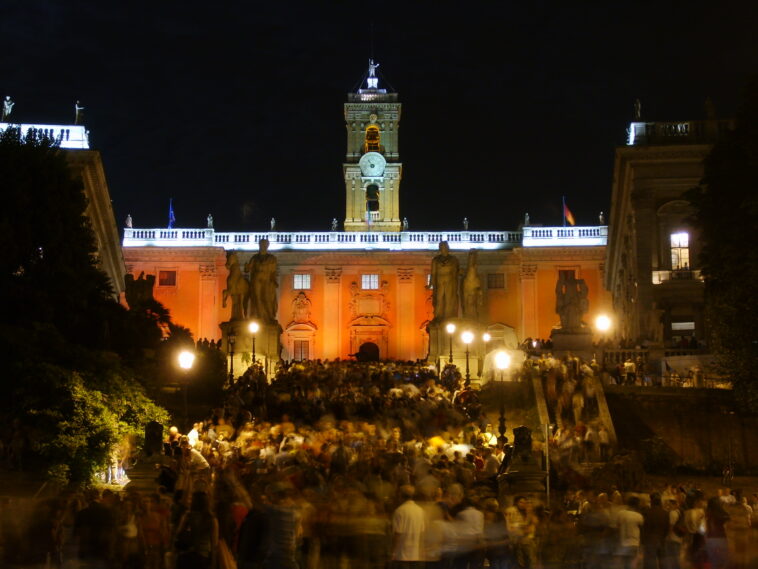 Piazza del Campidoglio illuminata durante un evento serale a Roma, con centinaia di persone riunite sulla scalinata e nella piazza davanti al Palazzo Senatorio.