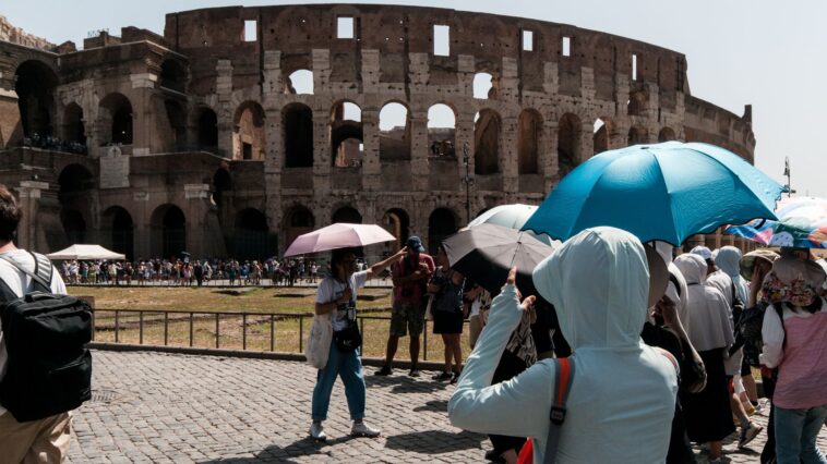 Turisti si proteggono dal sole con ombrelli davanti al Colosseo durante una giornata di forte caldo a Roma, mentre le temperature superano i 35 gradi.