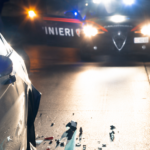 Auto danneggiata dopo un incidente stradale notturno, con detriti sull’asfalto e una pattuglia dei carabinieri sullo sfondo impegnata nei rilievi.