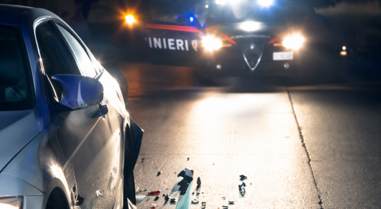 Auto danneggiata dopo un incidente stradale notturno, con detriti sull’asfalto e una pattuglia dei carabinieri sullo sfondo impegnata nei rilievi.