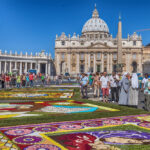 Piazza San Pietro decorata con i tradizionali tappeti floreali dell'Infiorata, mentre turisti, fedeli e religiose passeggiano tra le composizioni artistiche davanti alla Basilica di San Pietro.