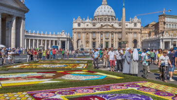 Piazza San Pietro decorata con i tradizionali tappeti floreali dell'Infiorata, mentre turisti, fedeli e religiose passeggiano tra le composizioni artistiche davanti alla Basilica di San Pietro.