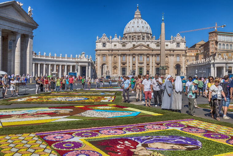 Piazza San Pietro decorata con i tradizionali tappeti floreali dell'Infiorata, mentre turisti, fedeli e religiose passeggiano tra le composizioni artistiche davanti alla Basilica di San Pietro.