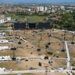 Veduta aerea dell’area di Tor Vergata durante i preparativi per il concerto di Ultimo, con il grande palco in fase di allestimento, tensostrutture, torri audio e luci, mezzi tecnici e lo skyline di Roma sullo sfondo.