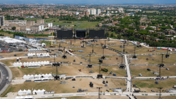 Veduta aerea dell’area di Tor Vergata durante i preparativi per il concerto di Ultimo, con il grande palco in fase di allestimento, tensostrutture, torri audio e luci, mezzi tecnici e lo skyline di Roma sullo sfondo.