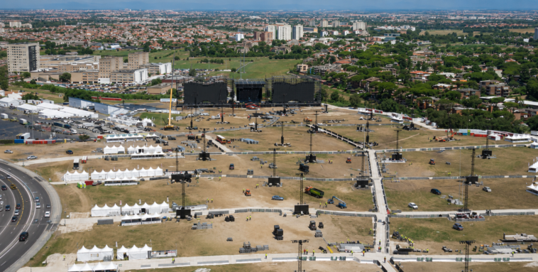 Veduta aerea dell’area di Tor Vergata durante i preparativi per il concerto di Ultimo, con il grande palco in fase di allestimento, tensostrutture, torri audio e luci, mezzi tecnici e lo skyline di Roma sullo sfondo.