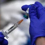 Un operatore sanitario con guanti blu prepara una dose di vaccino aspirando il liquido da una fiala con una siringa in un ambiente clinico.