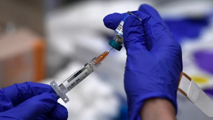 Un operatore sanitario con guanti blu prepara una dose di vaccino aspirando il liquido da una fiala con una siringa in un ambiente clinico.
