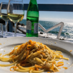 Piatto di spaghetti ai frutti di mare servito su una terrazza vista mare, con calici di vino bianco e il litorale sullo sfondo.
