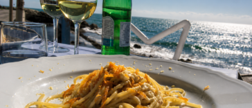 Piatto di spaghetti ai frutti di mare servito su una terrazza vista mare, con calici di vino bianco e il litorale sullo sfondo.