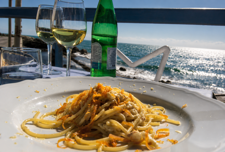 Piatto di spaghetti ai frutti di mare servito su una terrazza vista mare, con calici di vino bianco e il litorale sullo sfondo.