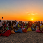 Persone sedute sui pouf colorati sulla spiaggia durante il tramonto, mentre il sole cala sul mare del litorale romano.