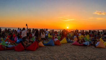 Persone sedute sui pouf colorati sulla spiaggia durante il tramonto, mentre il sole cala sul mare del litorale romano.