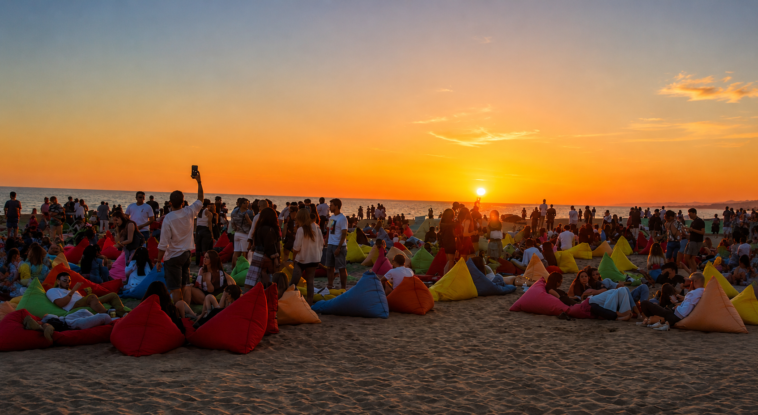 Persone sedute sui pouf colorati sulla spiaggia durante il tramonto, mentre il sole cala sul mare del litorale romano.