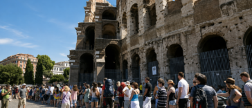 Turisti in fila davanti al Colosseo a Roma durante una giornata estiva, sotto il sole e con temperature elevate.
