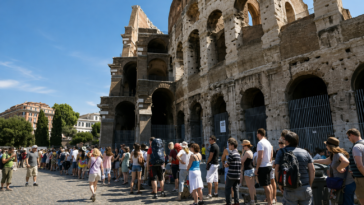 Turisti in fila davanti al Colosseo a Roma durante una giornata estiva, sotto il sole e con temperature elevate.