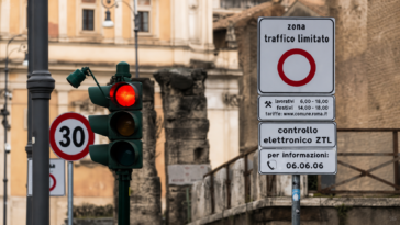Segnale della Ztl di Roma con controllo elettronico degli accessi e semaforo rosso, sullo sfondo edifici e resti storici del centro della Capitale.