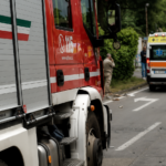 Mezzi di soccorso e ambulanza lungo una strada dopo un incidente, con detriti presenti sulla carreggiata.