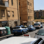Strada nel quartiere Alessandrino a Roma con diverse auto parcheggiate e mezzi della polizia presenti vicino ad alcuni palazzi.