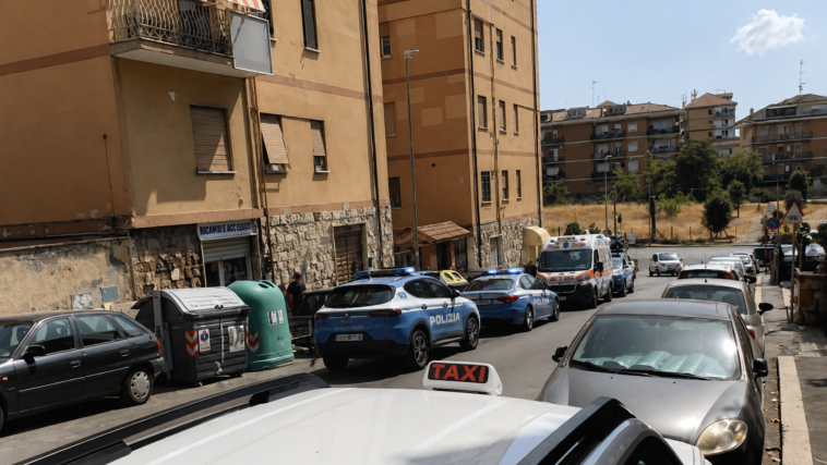 Strada nel quartiere Alessandrino a Roma con diverse auto parcheggiate e mezzi della polizia presenti vicino ad alcuni palazzi.