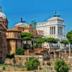 Vista dei Fori Imperiali a Roma con resti archeologici, chiese storiche, alberi e il Vittoriano sullo sfondo.