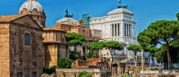 Vista dei Fori Imperiali a Roma con resti archeologici, chiese storiche, alberi e il Vittoriano sullo sfondo.