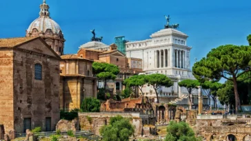 Vista dei Fori Imperiali a Roma con resti archeologici, chiese storiche, alberi e il Vittoriano sullo sfondo.
