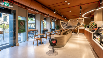 Interno di una moderna caffetteria con tavoli, sedute, ampie vetrate e bancone in legno.